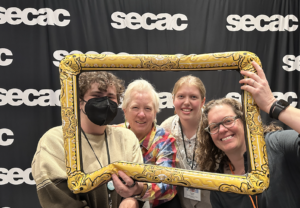 (l-r) August Fisk '25, Nell Ruby, Tallulah Stroud '26, Katherine Smith in front of secac step and repeat photo banner holding an inflatable gilded frame