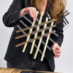Jess Jones holds up a simple weaving of three colors of paper strips laid in three directions on a table top