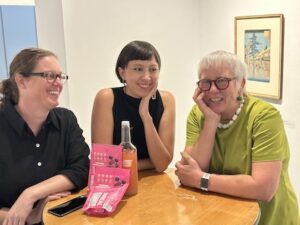 co-curators Katherine Smith, Anna Carnes and Nell Ruby smiling together at a table during the opening of the Reading Room 