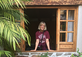 Image of Susan Meyers, shoulder length hair, smiling, looking out of a large open window, arms in front of her resting on the sill